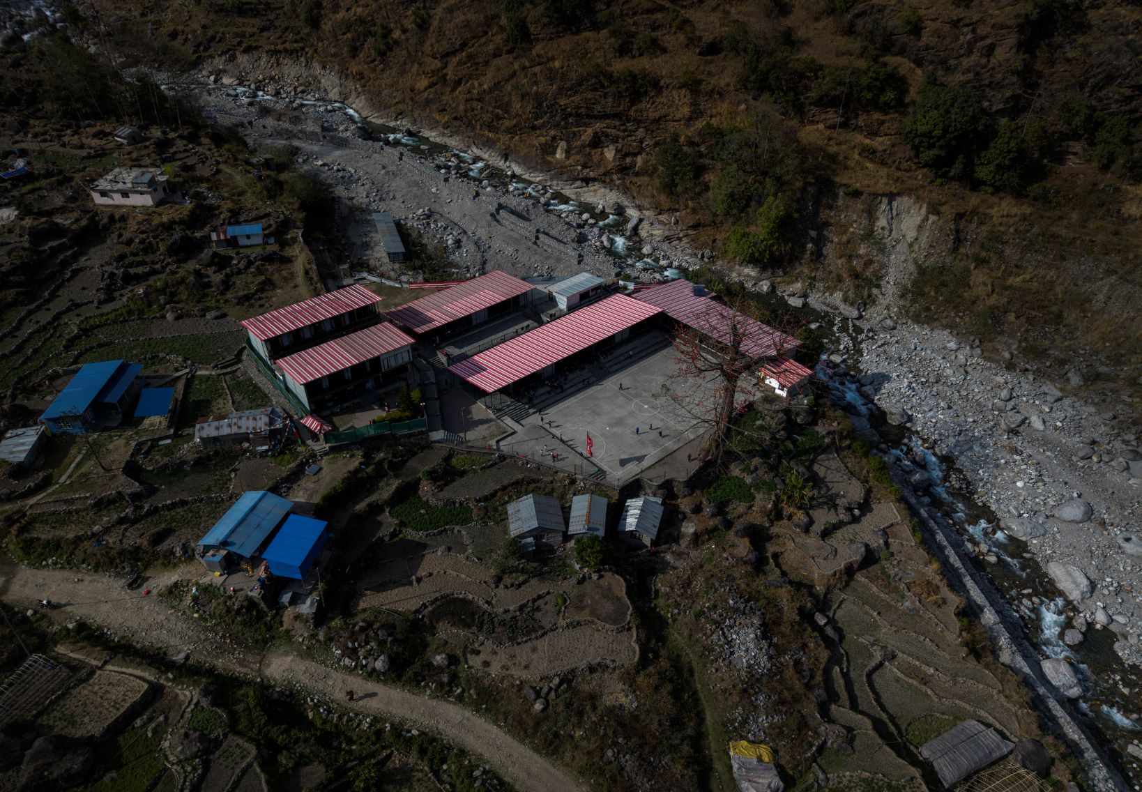 Aerial view of a rural school with red roofs bordered by rocky terrain, farmlands, small homes, and a riverbed.