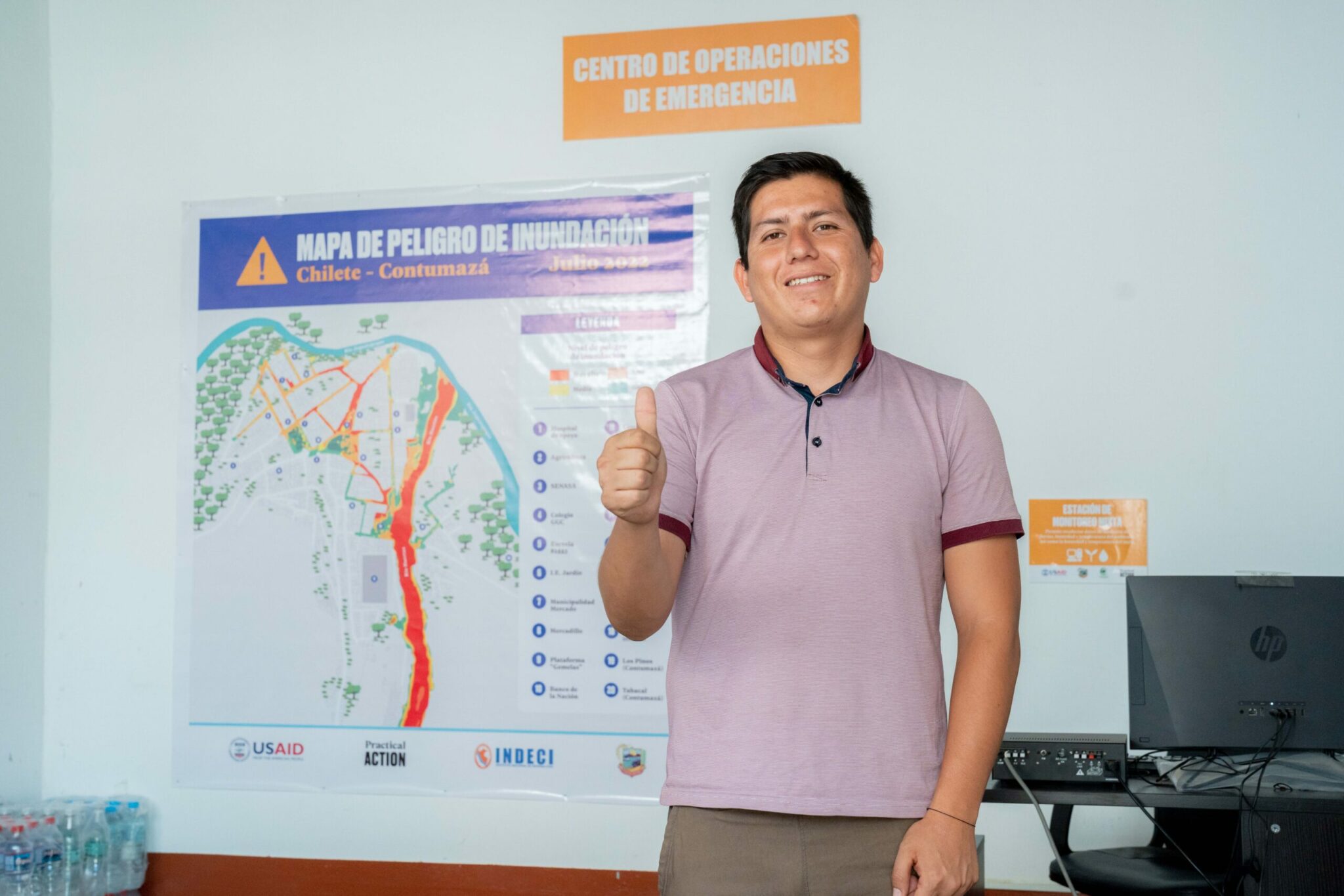 A man stands indoors giving a thumbs-up in front of a flood hazard map and an emergency operations center sign.