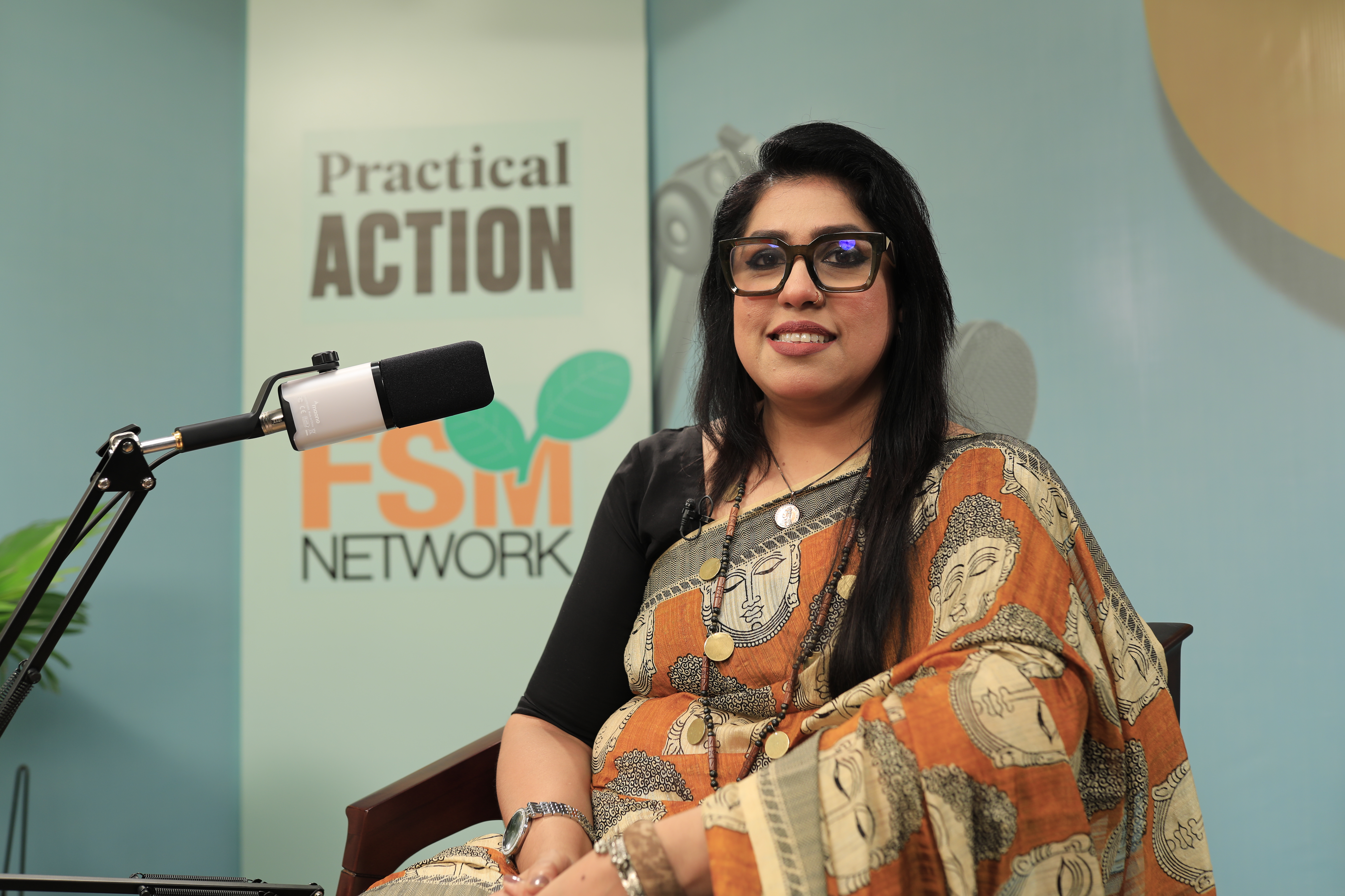 A woman wearing glasses and a printed saree sits in front of a microphone, with 