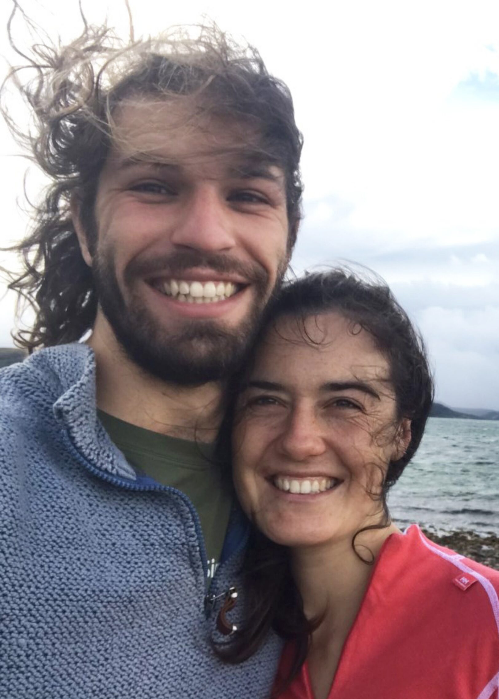 Two people smiling and standing close together outdoors near a body of water on a windy day.