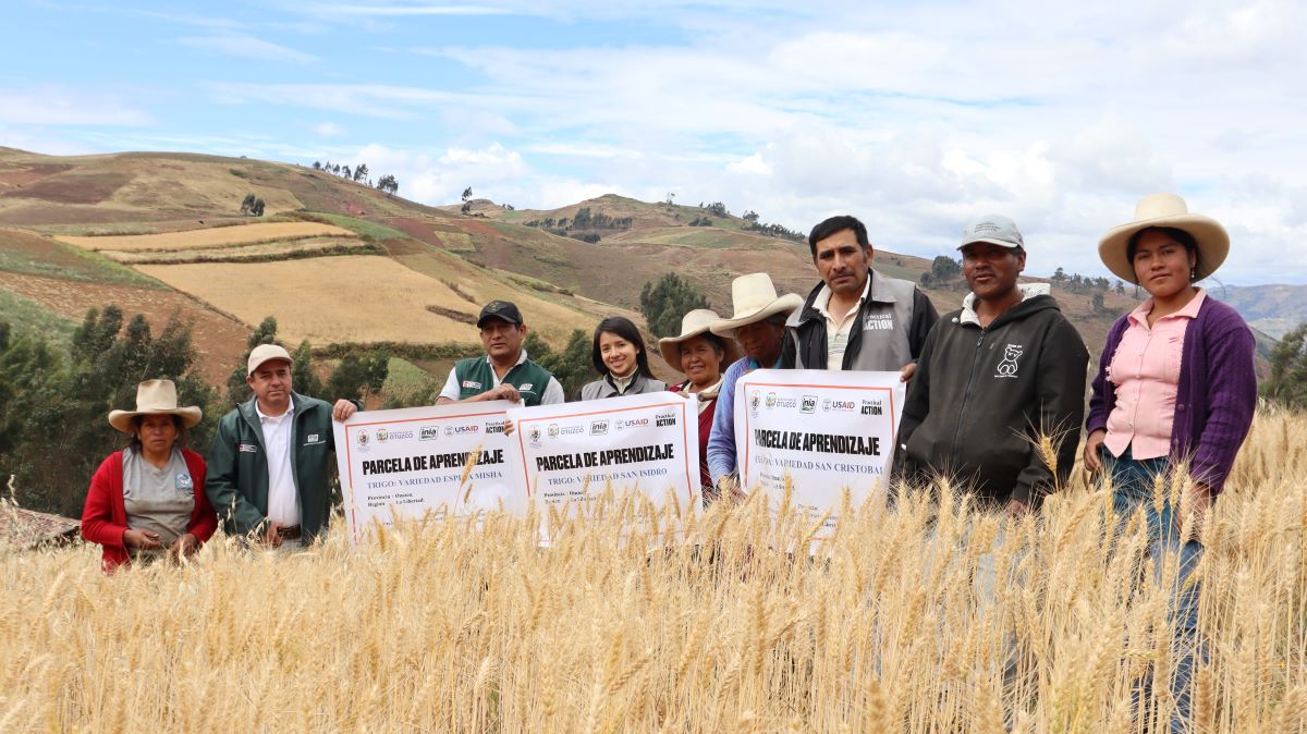 A group of people stands in a wheat field holding signs with text, with hills and farmland visible in the background under a partly cloudy sky.
