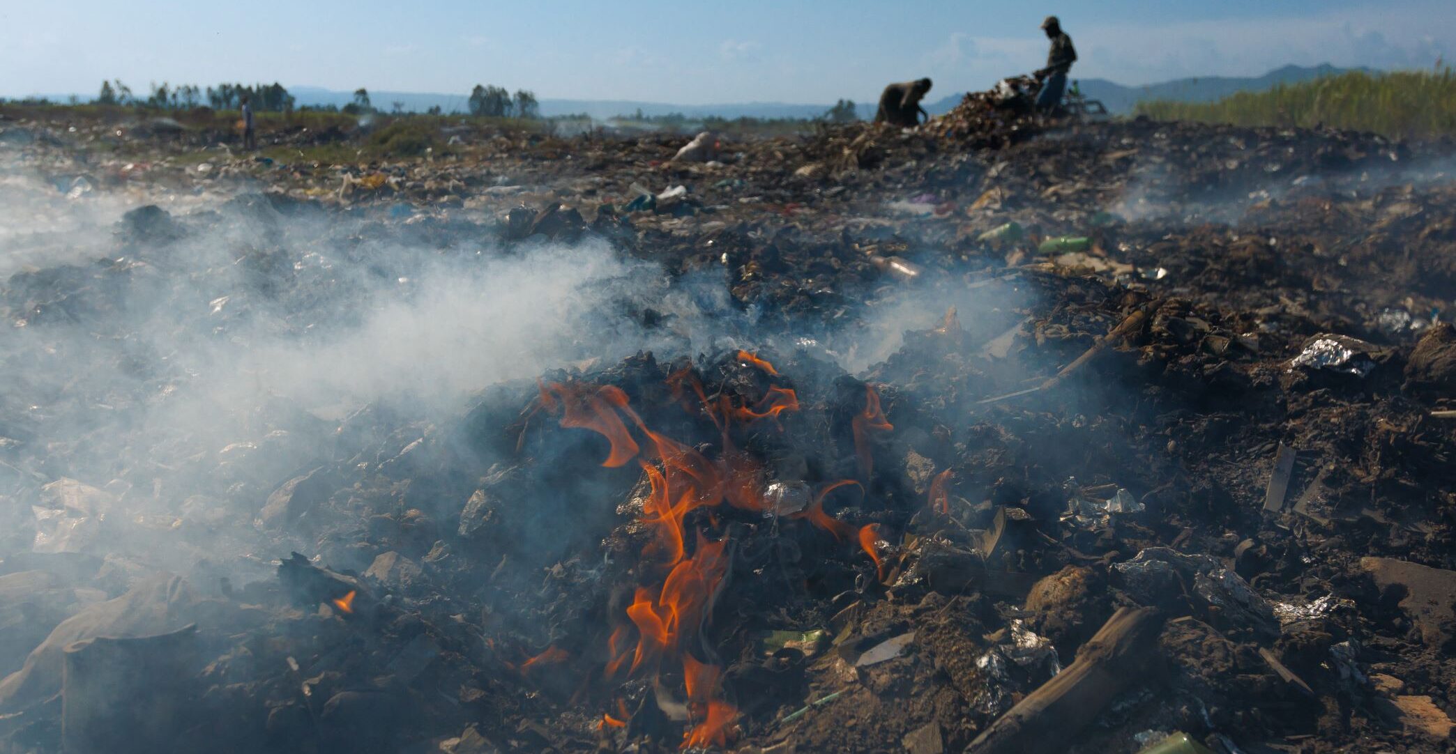 A fire burns amid piles of garbage in an open landfill with smoke rising and people working in the background under a clear sky.