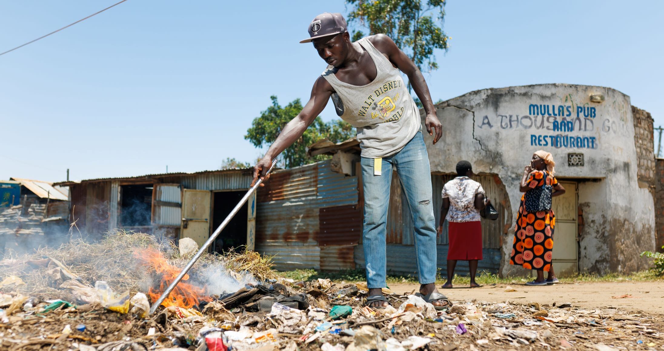 A man uses a stick to tend to a pile of burning trash by the roadside near a building labeled "Mullas Pub and Restaurant." Two women stand outside the building in the background.