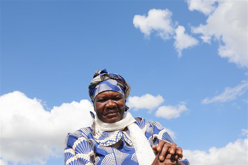 An older woman dressed in a blue and white patterned outfit and headscarf stands against a backdrop of blue sky with scattered clouds.