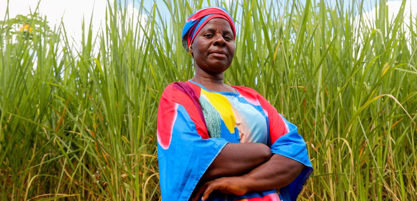 Woman in a colorful dress and headwrap stands with arms crossed in front of tall green grass under a partly cloudy sky.