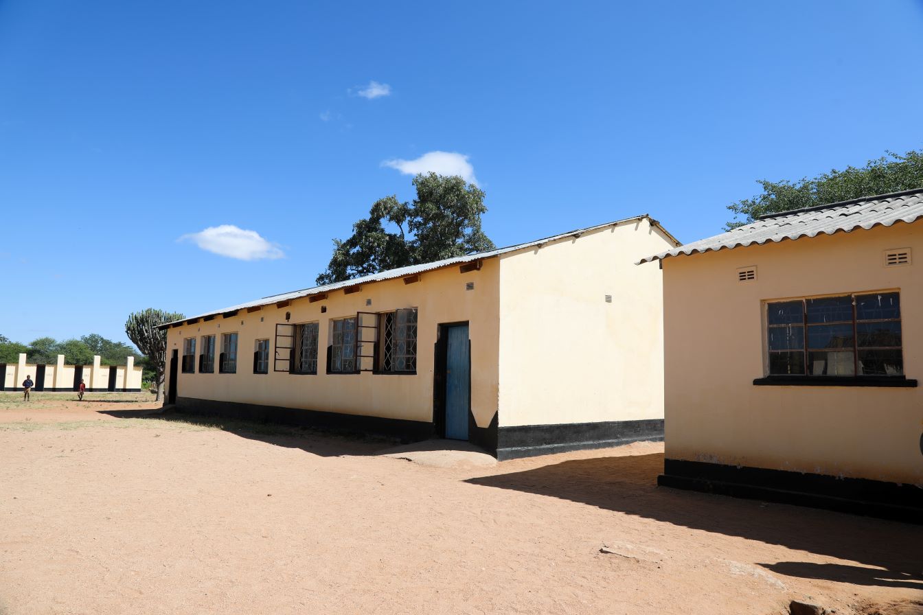 Single-story yellow school buildings with tin roofs stand on a sandy yard under a clear blue sky, with trees in the background.