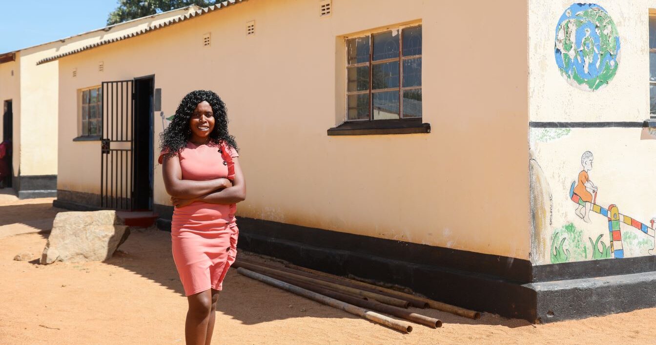 A woman in a coral dress stands with arms crossed in front of a cream-colored building with painted murals and barred windows on a sunny day.