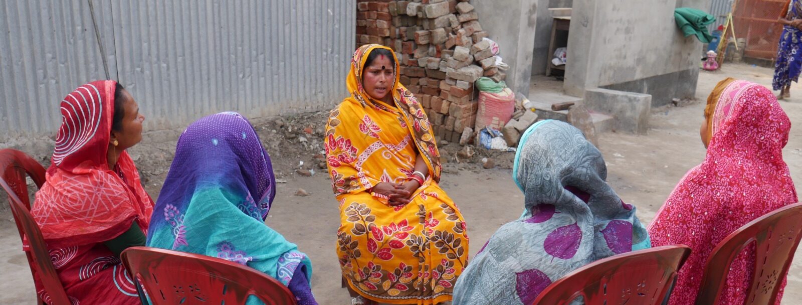 Six women in colorful sarees sit in a circle on red plastic chairs outdoors, having a conversation near a corrugated metal and concrete building.