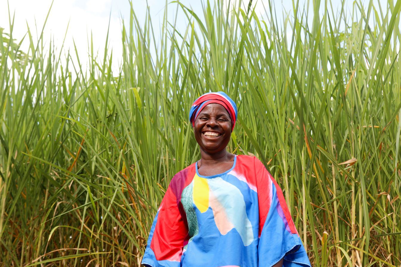 A woman in a colorful dress and headscarf stands smiling in front of tall green grass.