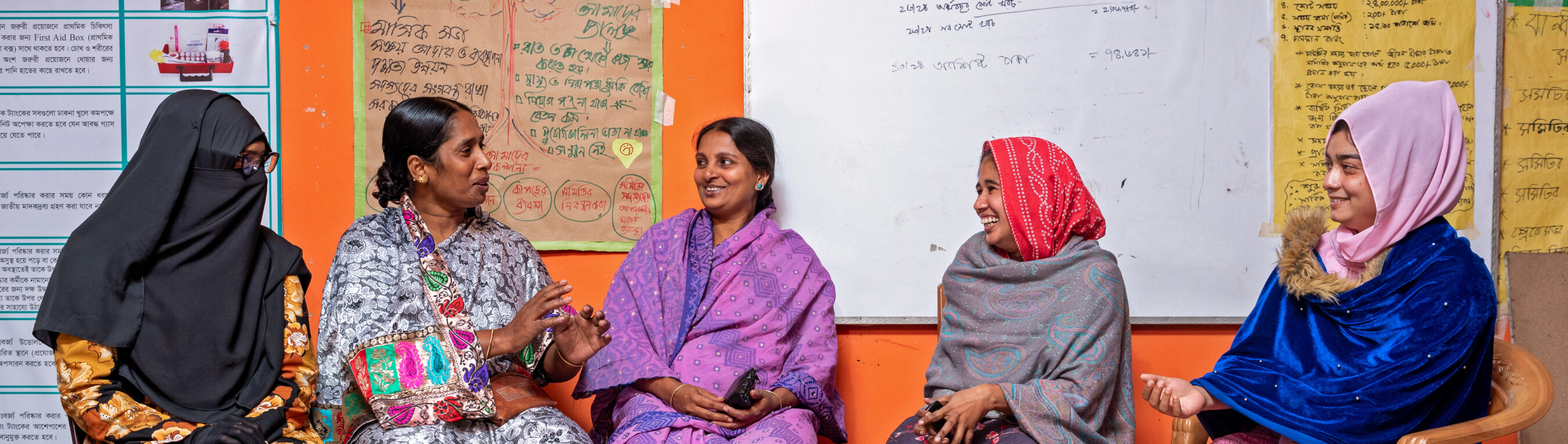 Five women sit together on plastic chairs, engaged in conversation, in front of colorful posters and whiteboards with writing in a classroom or community center setting.