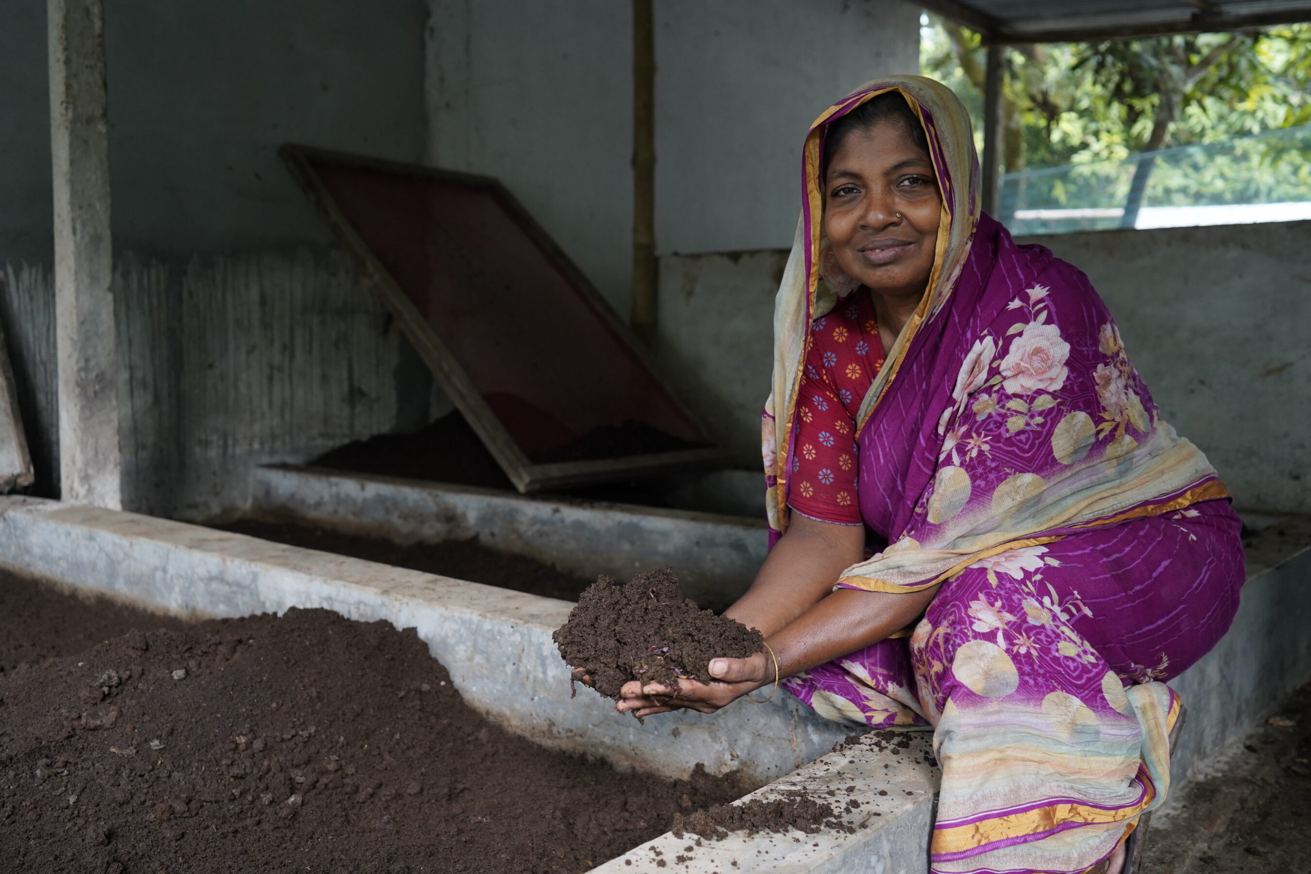 A woman in a colorful sari holds a handful of soil or compost, kneeling by large bins filled with soil in an indoor setting.