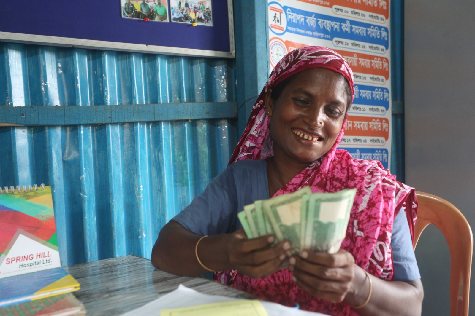 A woman in a pink patterned headscarf sits at a desk, smiling and counting paper currency bills in her hands.