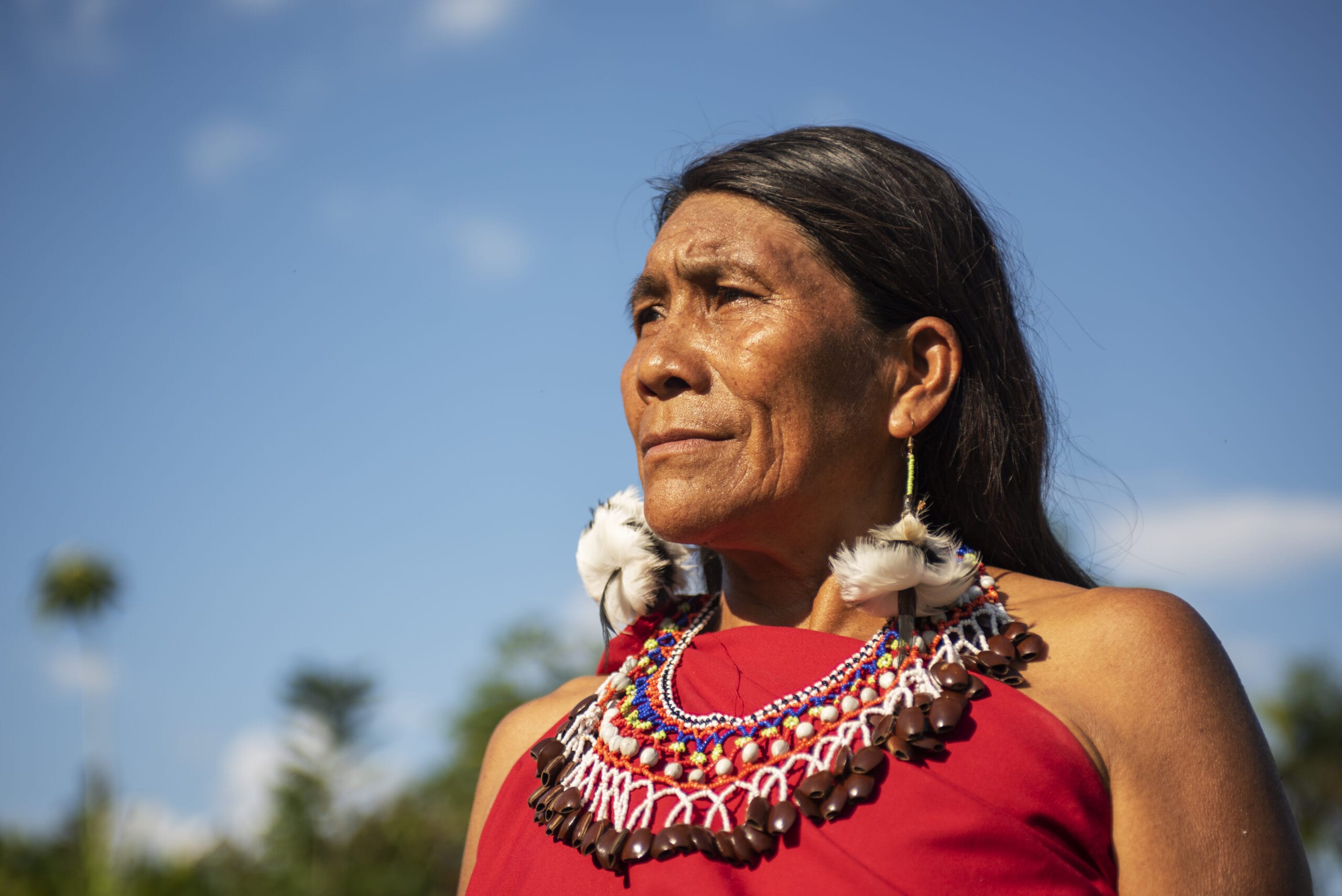 An Indigenous person with long dark hair wears a red garment, colorful beaded necklace, and earrings with feathers, standing outdoors against a blue sky.