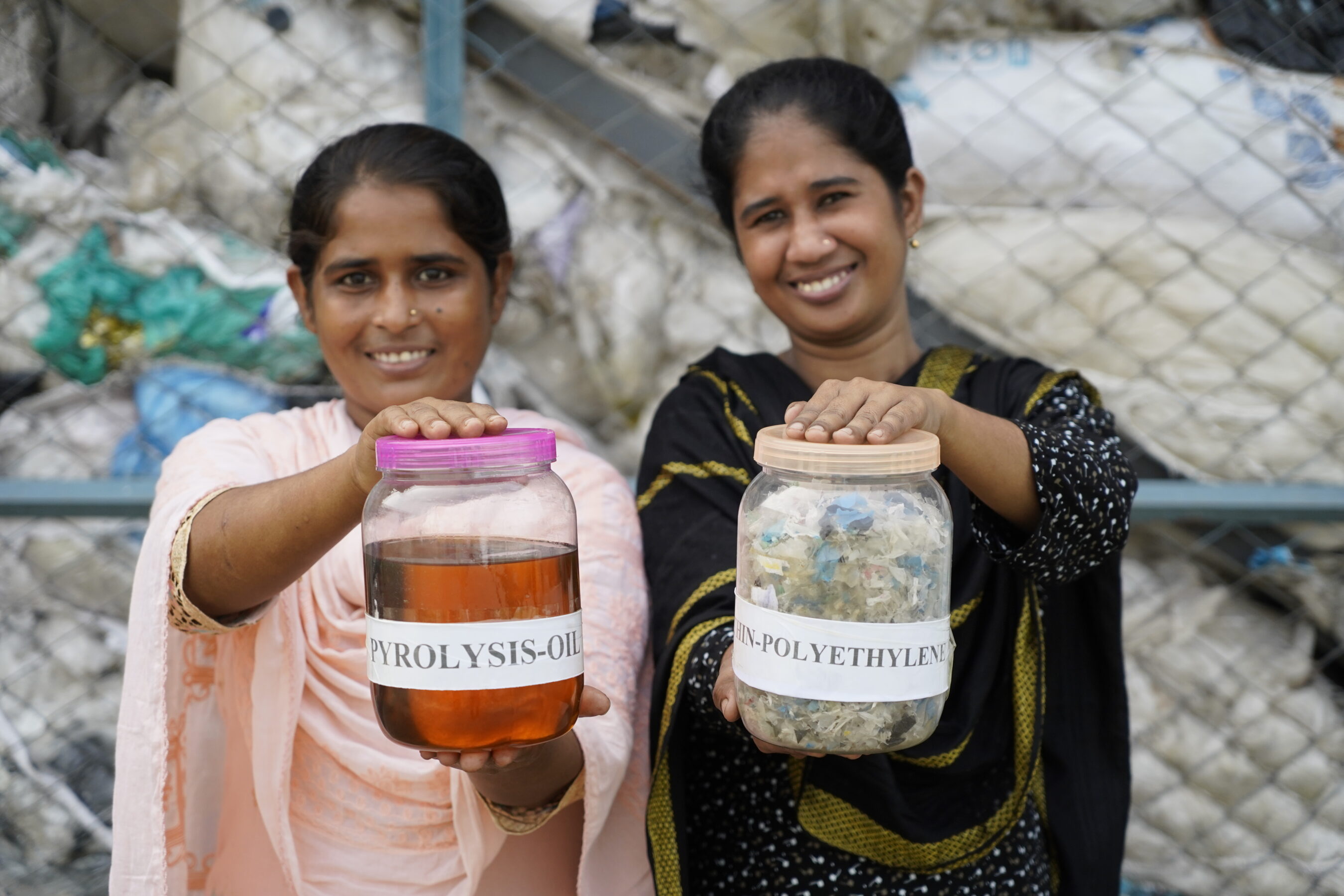Two women smile while holding jars labeled "Pyrolysis Oil" and "Polyethylene," standing in front of a fence with plastic waste in the background.