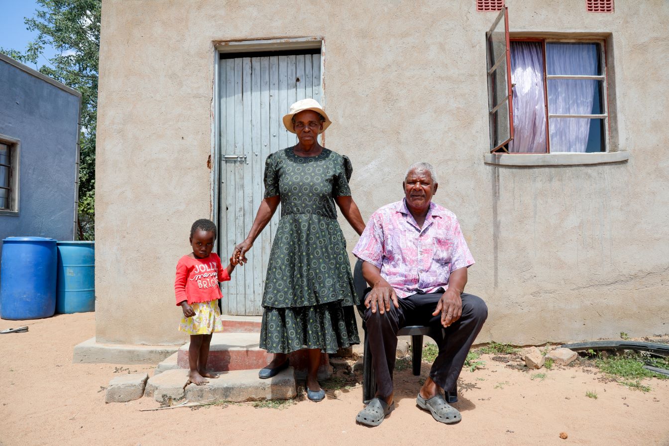 An older man sits on a chair next to an older woman standing with a young child outside a beige house with a wooden door and an open window.