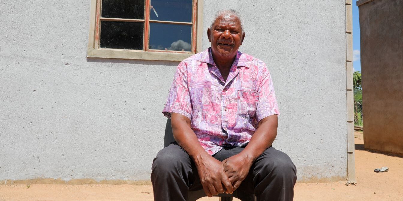 An older man in a pink patterned shirt sits on a chair in front of a gray concrete wall with a window, outdoors on a sunny day.