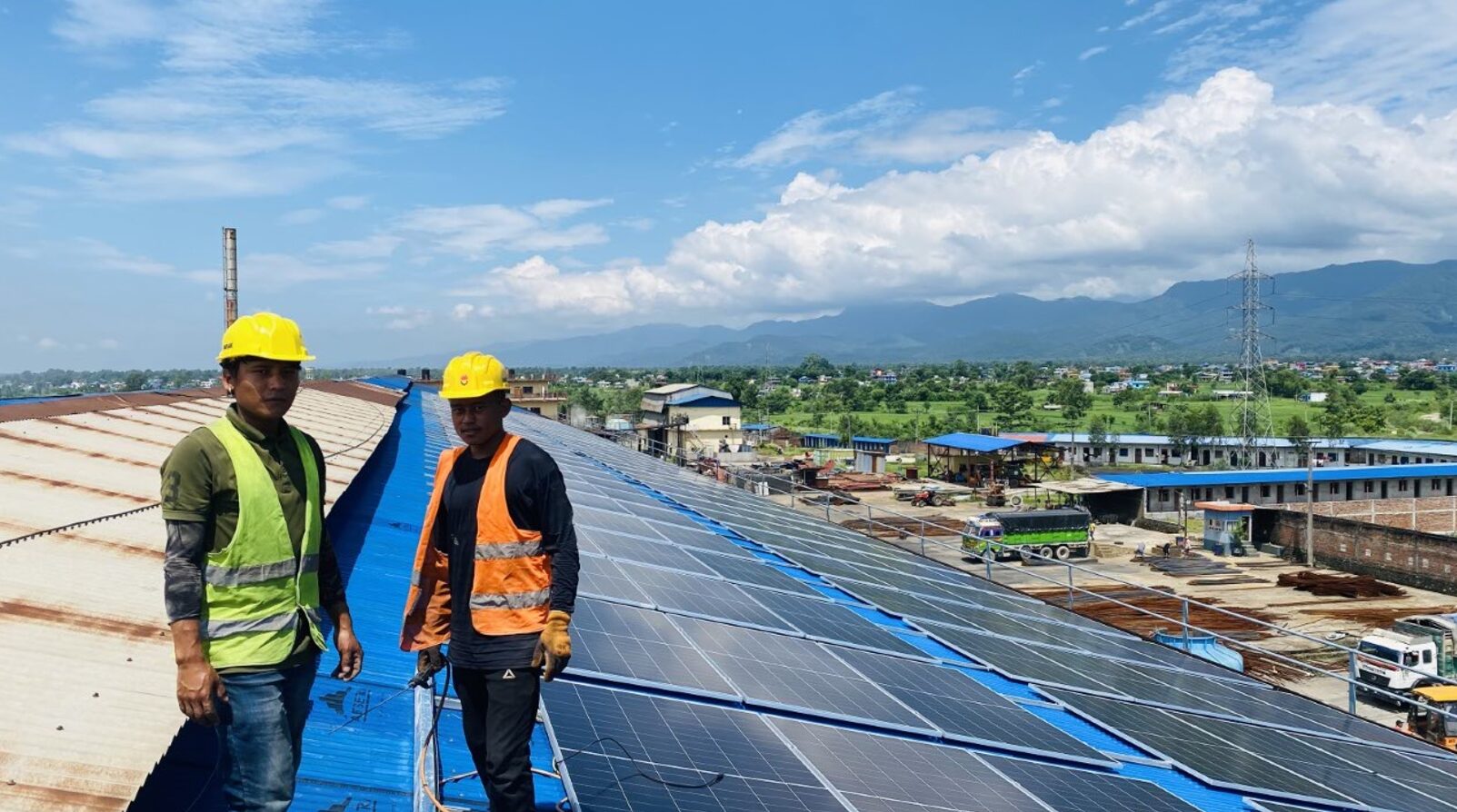 Two workers in safety gear stand on a rooftop with solar panels, overlooking an industrial area with mountains and clouds in the background.