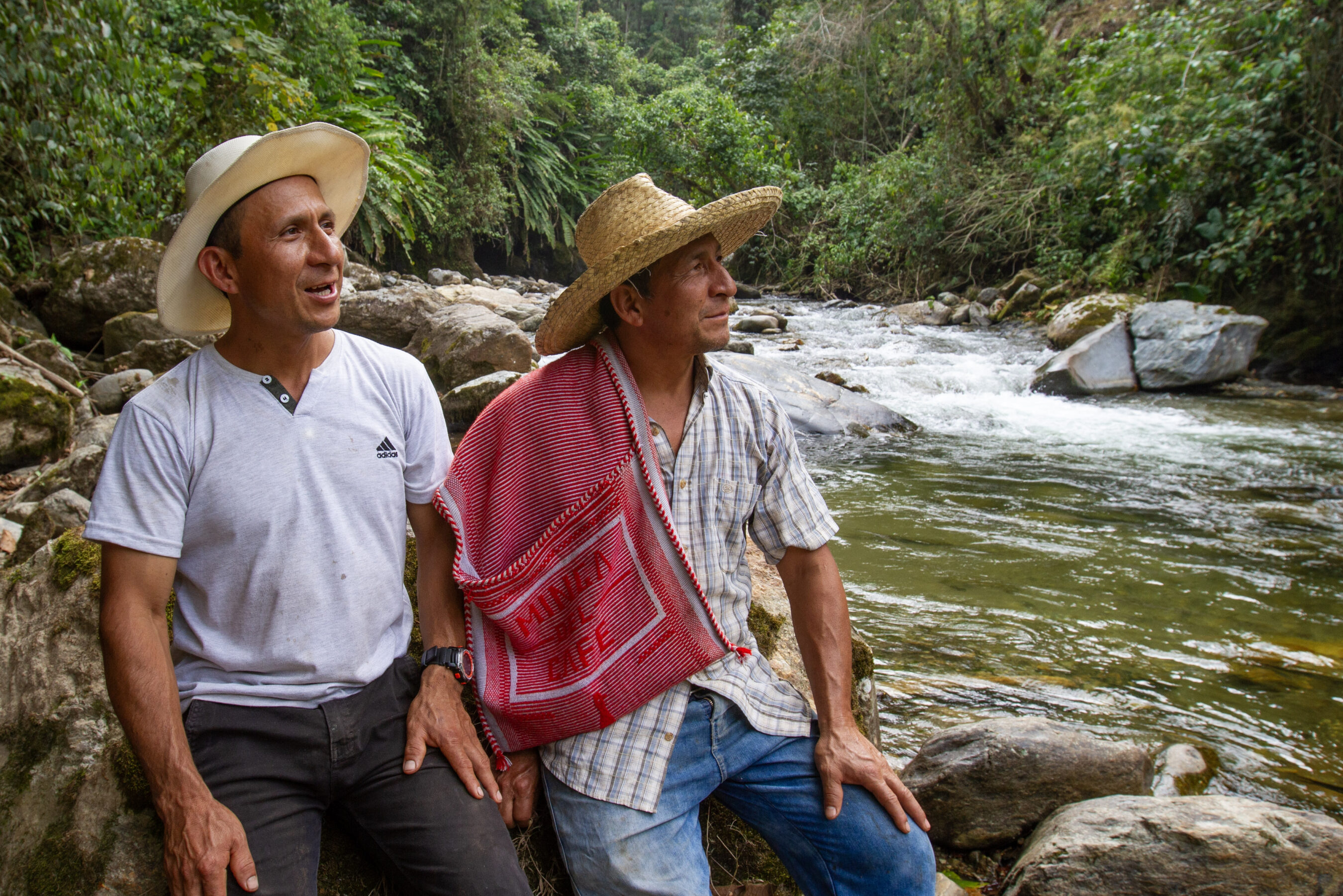 Two men wearing hats sit on rocks beside a river in a lush, green forest. One wears a white shirt, the other a plaid shirt with a red cloth draped over his shoulder.