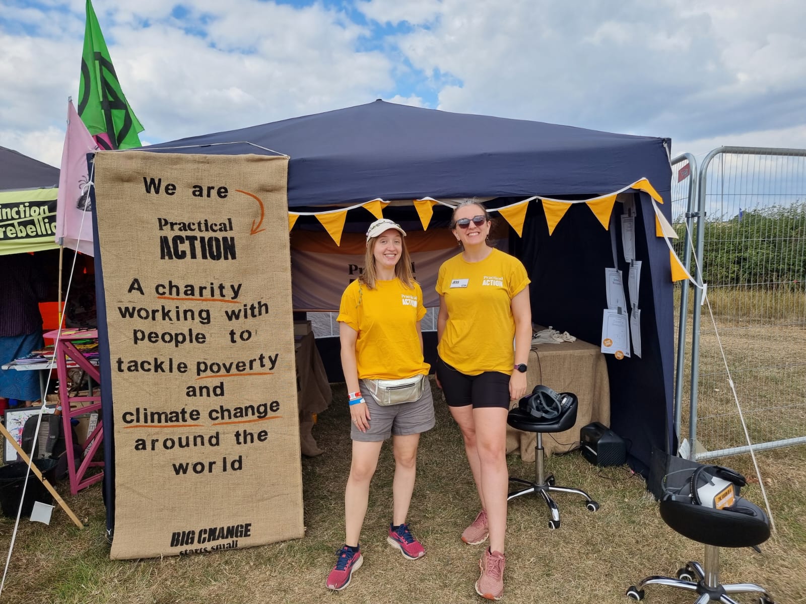 Two women in yellow shirts stand in front of a tent with a "Practical Action" banner about fighting poverty and climate change at an outdoor event.