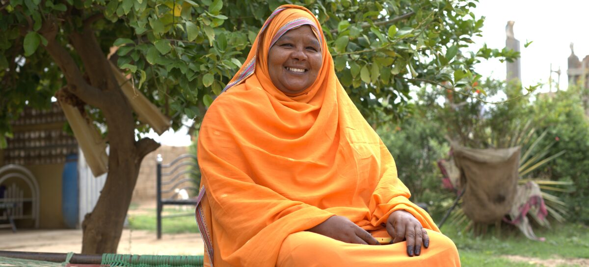 A woman wearing an orange hijab and dress sits outdoors on a woven bench, smiling, with greenery and trees in the background.