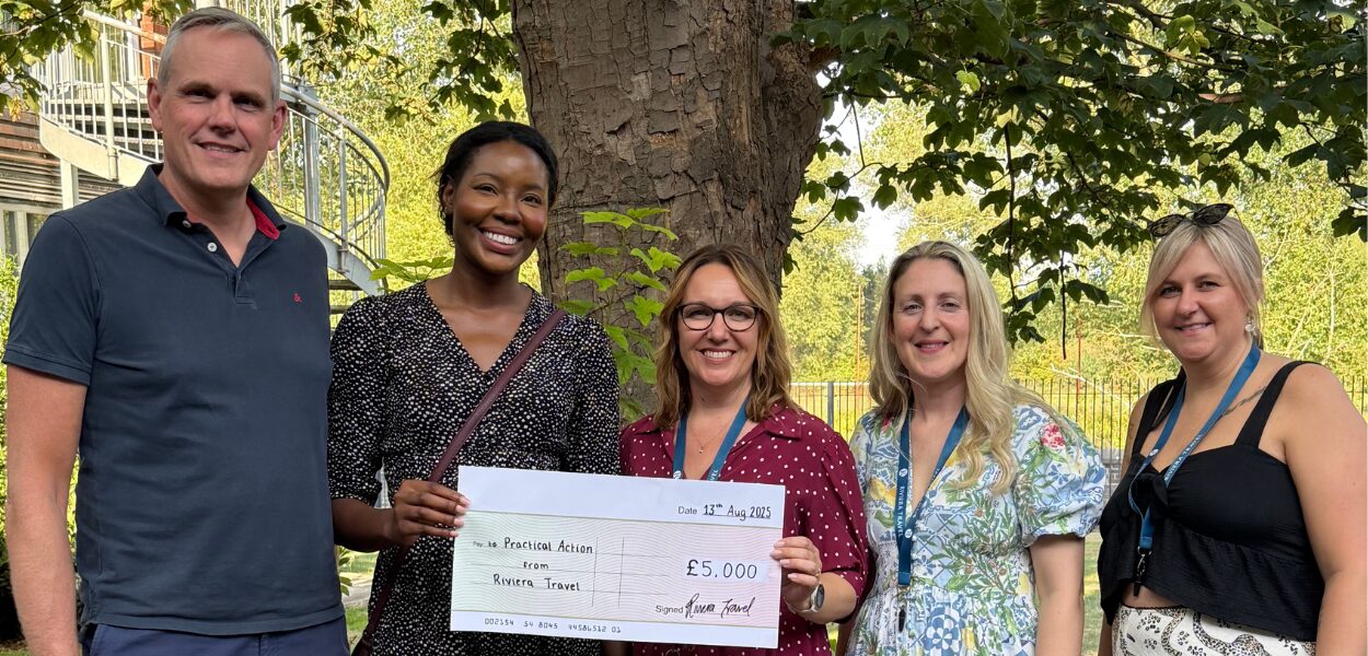 Five people stand outdoors, smiling and holding a large ceremonial check for £5,000 made out to "Firstline Admin" from "Rivers Trust.