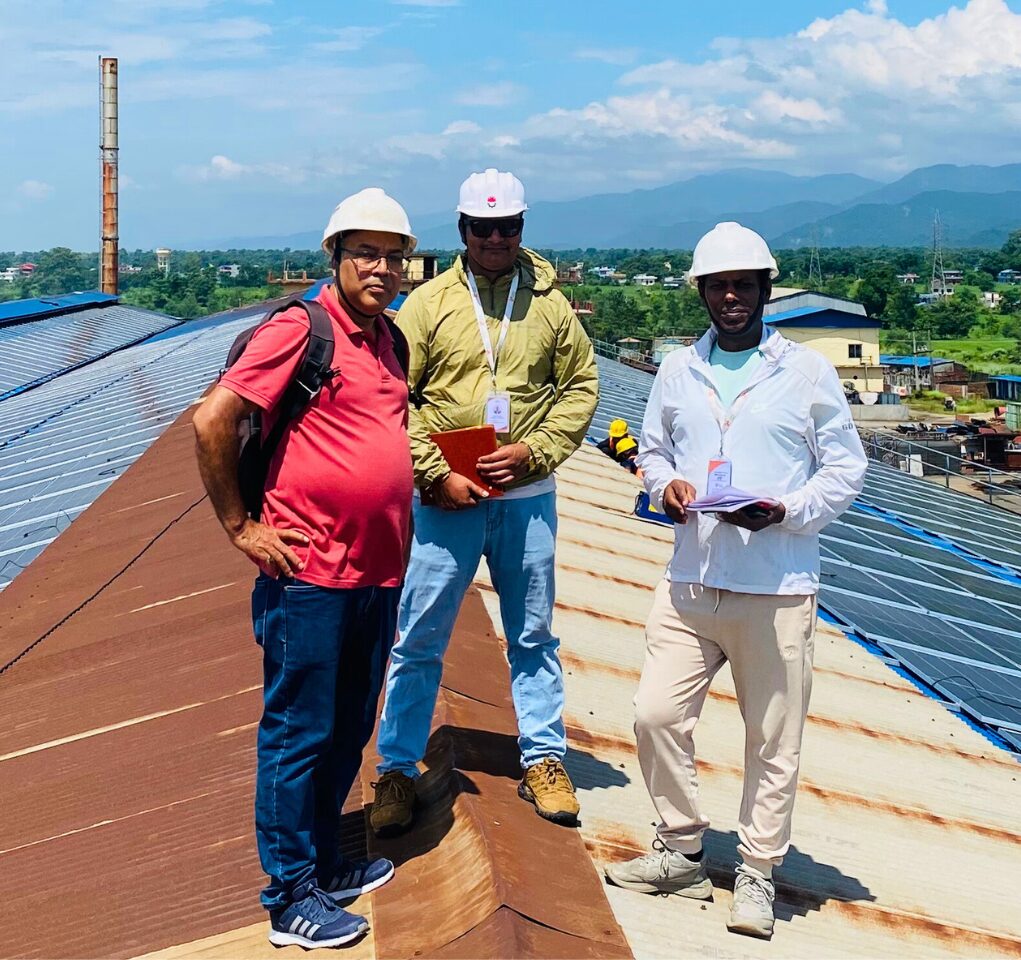 Three men wearing safety helmets and casual clothing stand on a sloped metal rooftop, holding notebooks, with industrial buildings and hills visible in the background.