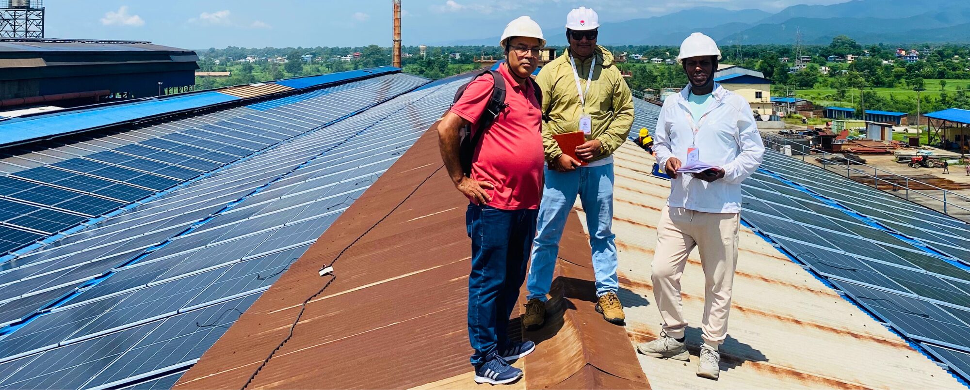 Three men in hard hats and safety gear stand on a rooftop covered with solar panels, with buildings and mountains visible in the background.