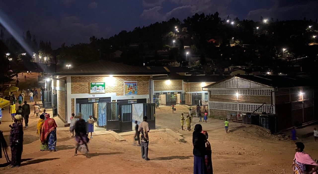 People walk and gather around a well-lit building complex at night, with hills and scattered lights visible in the background.