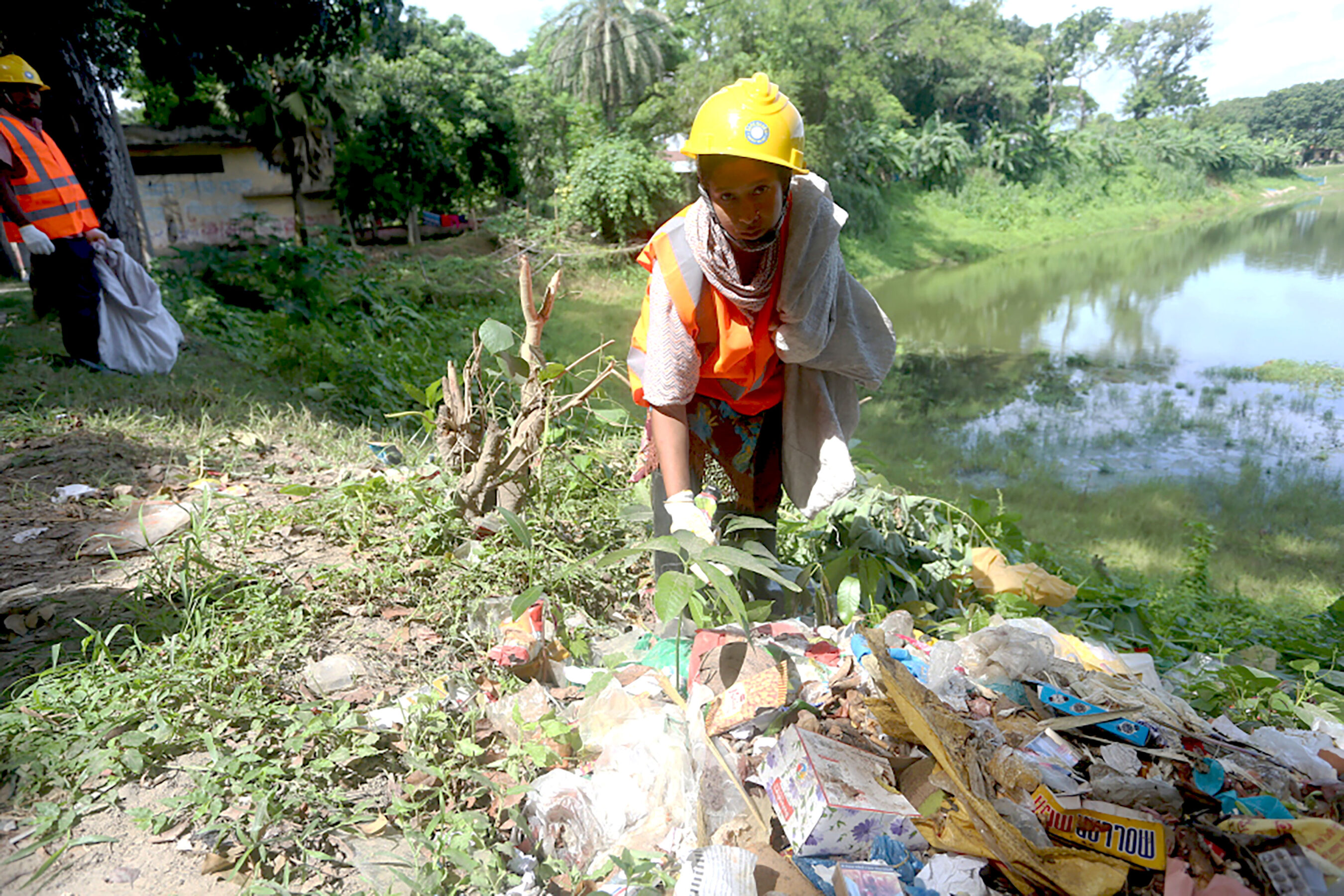 A worker in a safety vest and helmet collects trash near a riverbank, with scattered litter and greenery visible around.