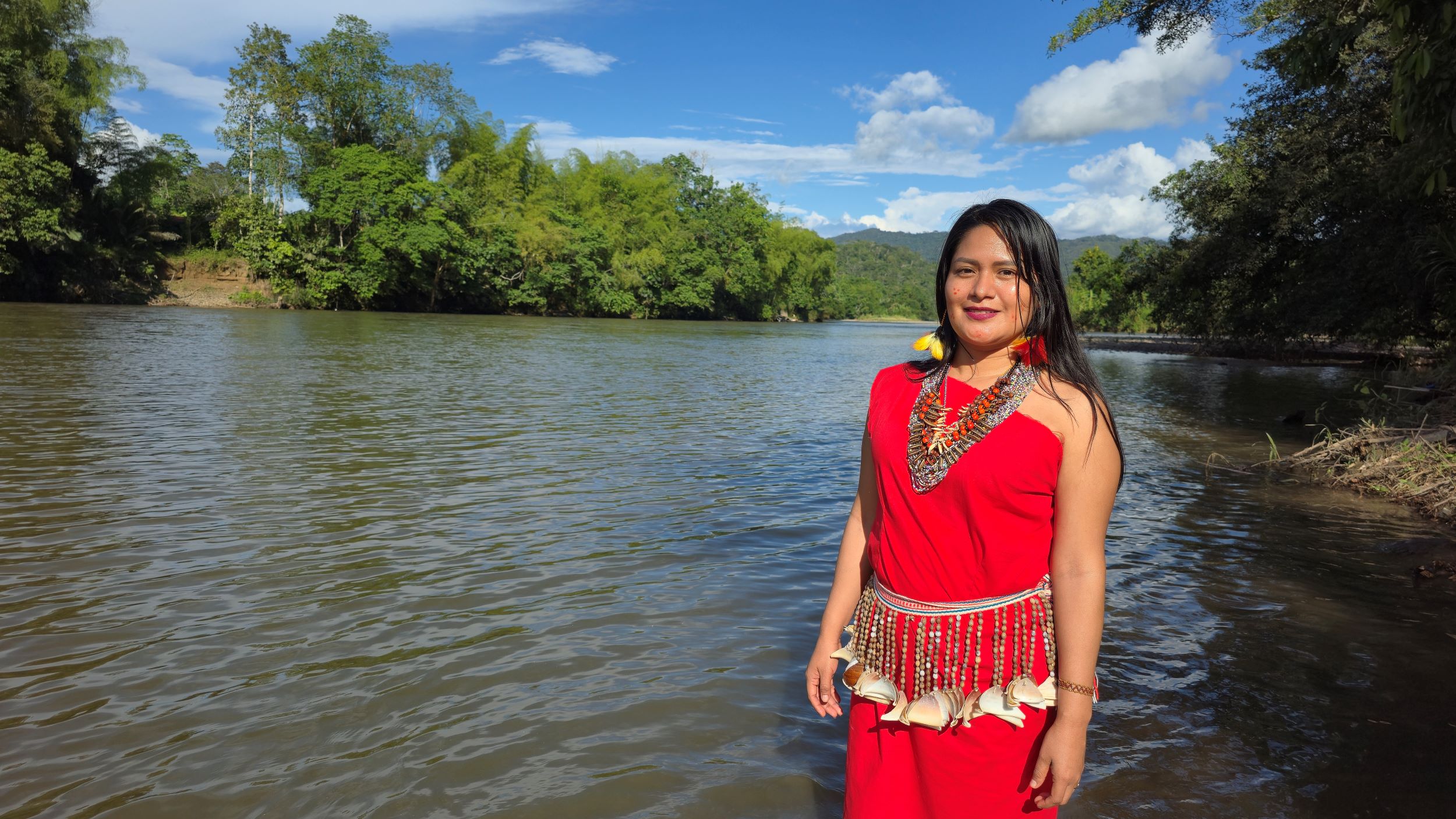A woman in a red dress with traditional jewelry stands by a riverbank surrounded by lush green trees under a blue sky.