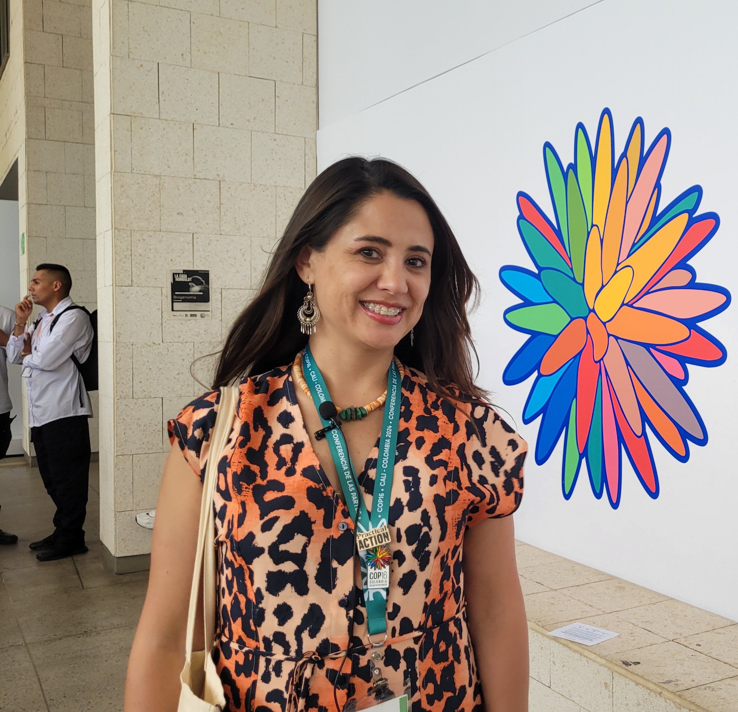 Woman wearing a leopard print dress and conference lanyard stands indoors near a colorful, abstract flower mural with other people in the background.