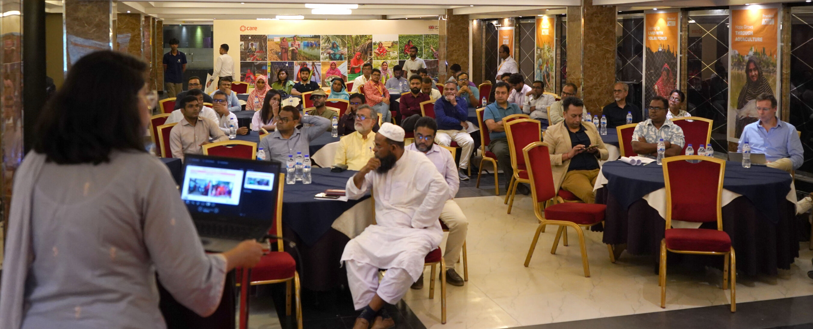 A woman presents to an audience seated at round tables in a conference room; people listen, take notes, or use devices.