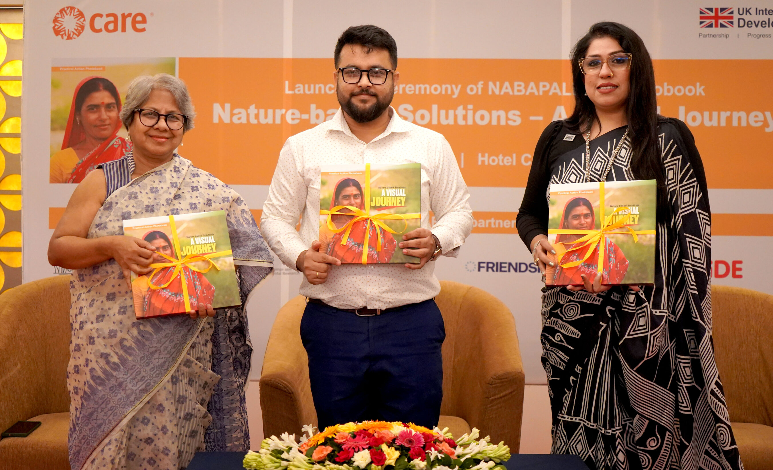 Three individuals stand side by side, each holding a book wrapped with a yellow ribbon, at a launch event with a “Nature-based Solutions” banner in the background.