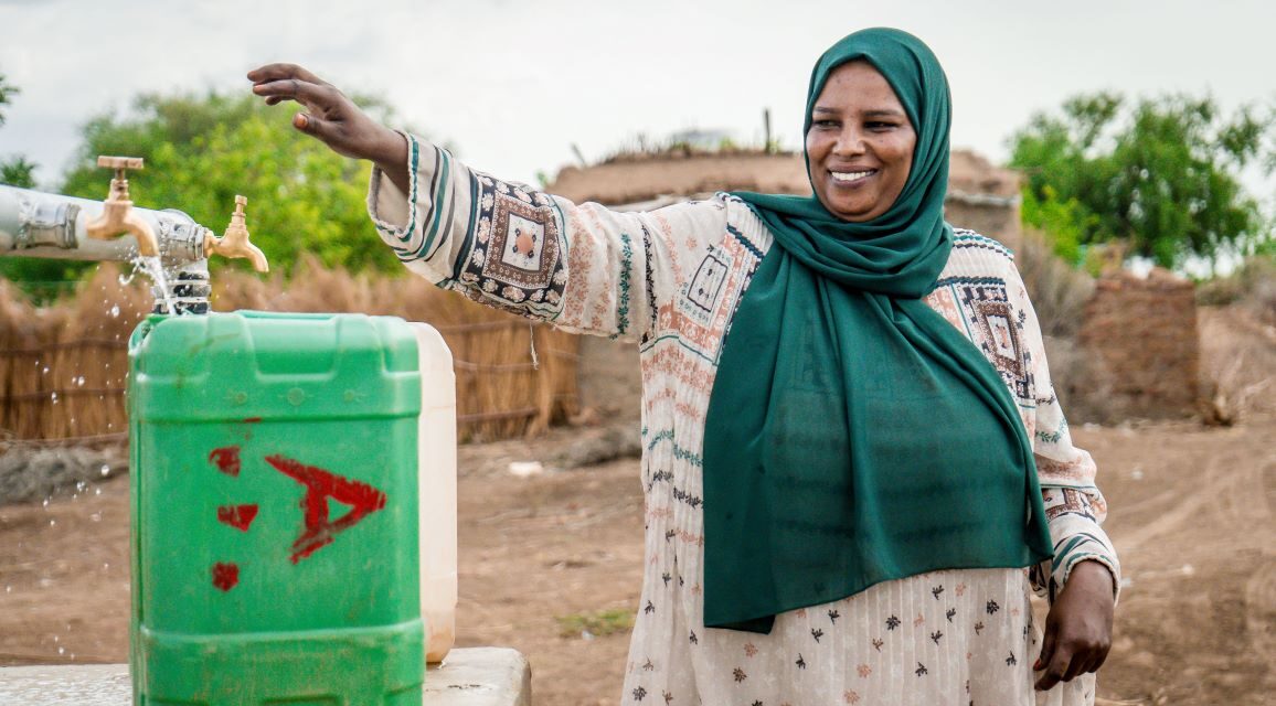 A woman in a green hijab smiles while filling a green jerrycan with water from an outdoor tap in a rural area.