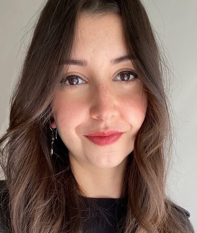 A woman with long brown hair and brown eyes looks at the camera, smiling slightly. She is wearing a black top and silver earrings. The background is plain and light-colored.