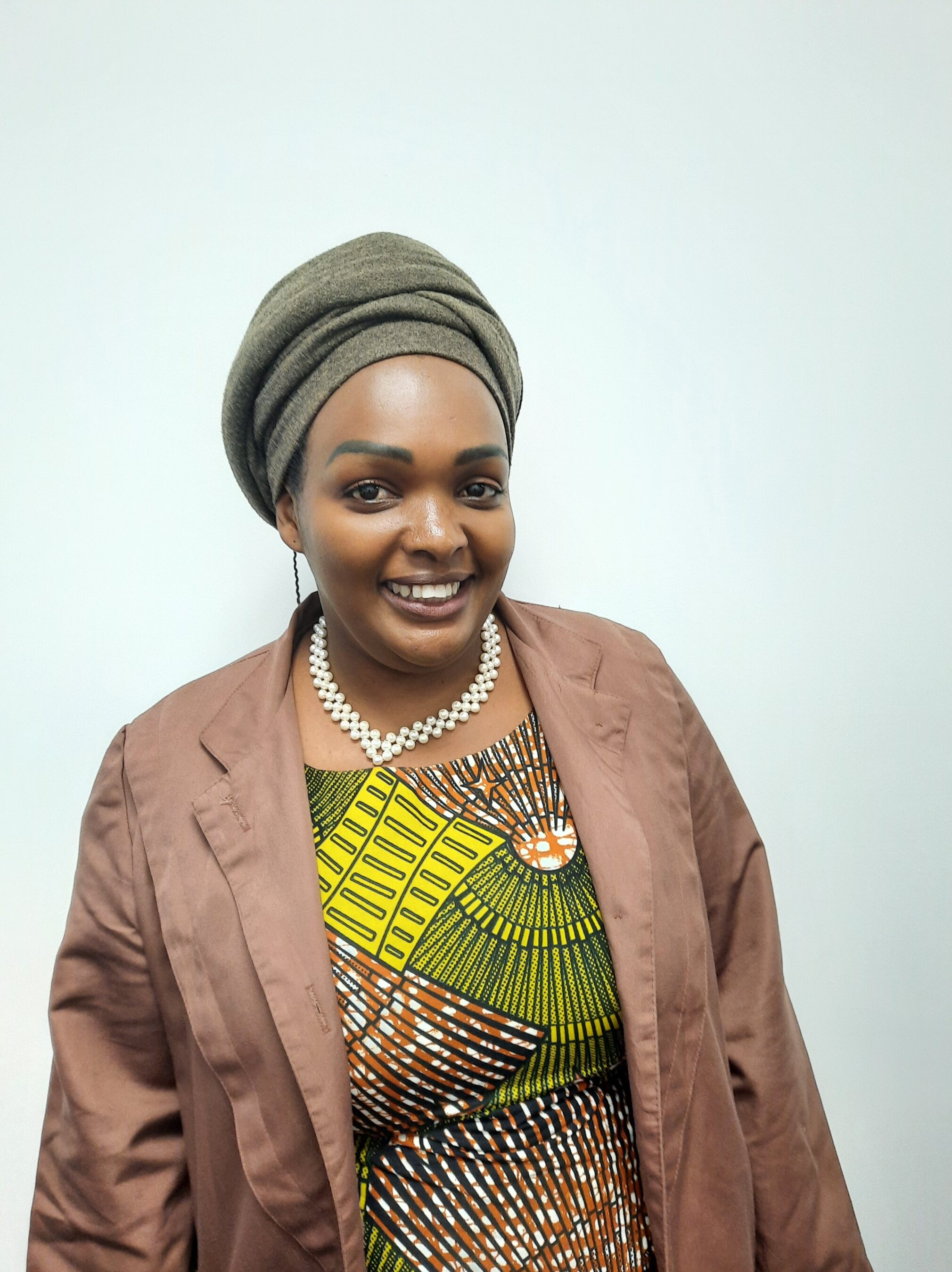 A woman wearing a headwrap, brown blazer, patterned dress, and pearl necklace smiles in front of a plain white background.