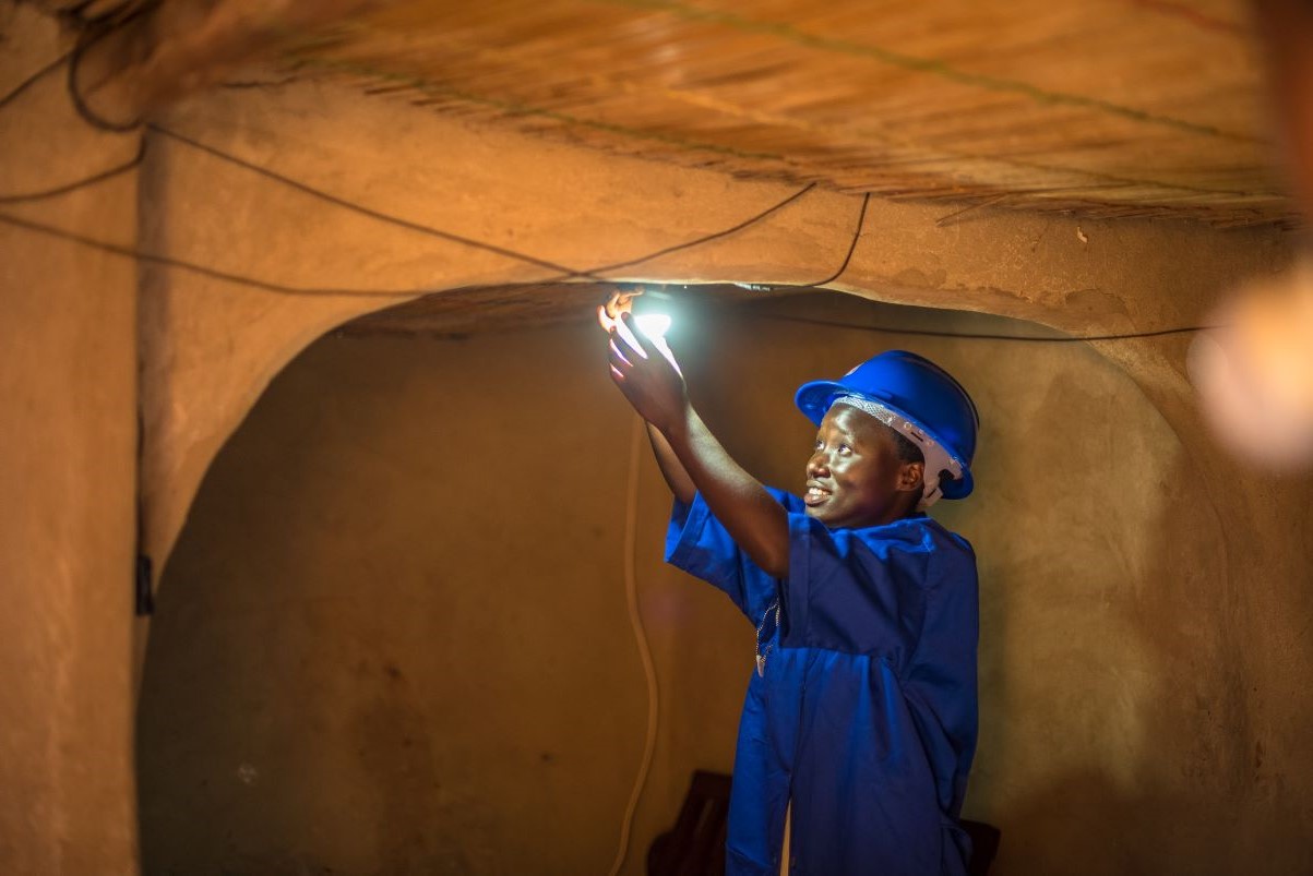 A person wearing a blue hard hat and shirt installs or repairs a light bulb on the ceiling in a dimly lit room with clay walls.
