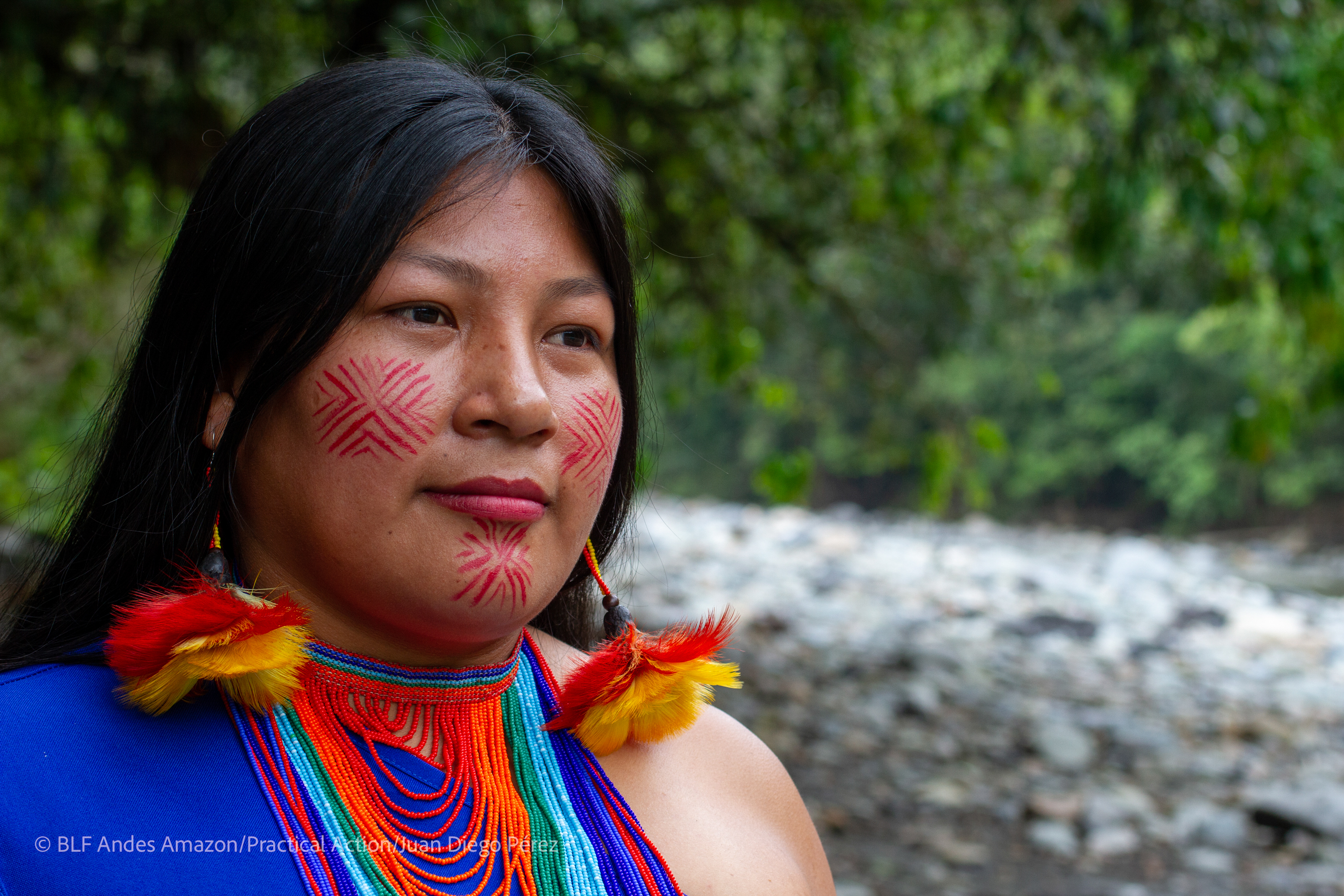 A woman with long dark hair, red geometric face paint, vibrant feather earrings, and a beaded necklace stands outdoors near a river and forest.