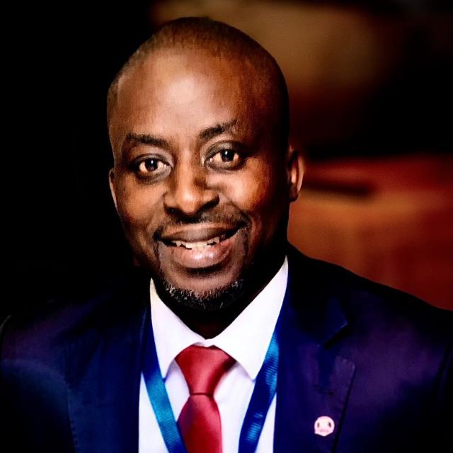 A man in a dark suit, white shirt, and red tie smiles at the camera. He has a short beard and wears a blue lanyard. The background is blurred.