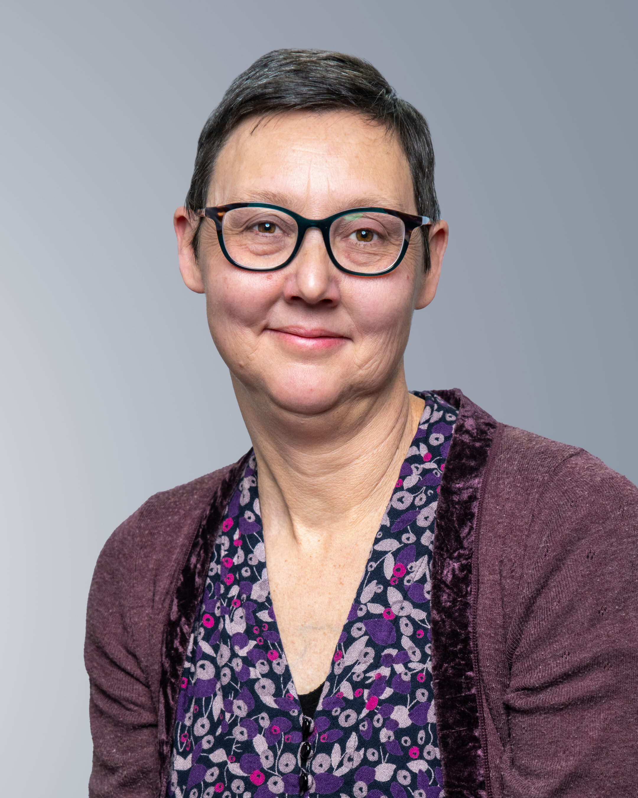 A woman with short dark hair and glasses, wearing a purple patterned blouse and maroon cardigan, poses against a plain gray background.