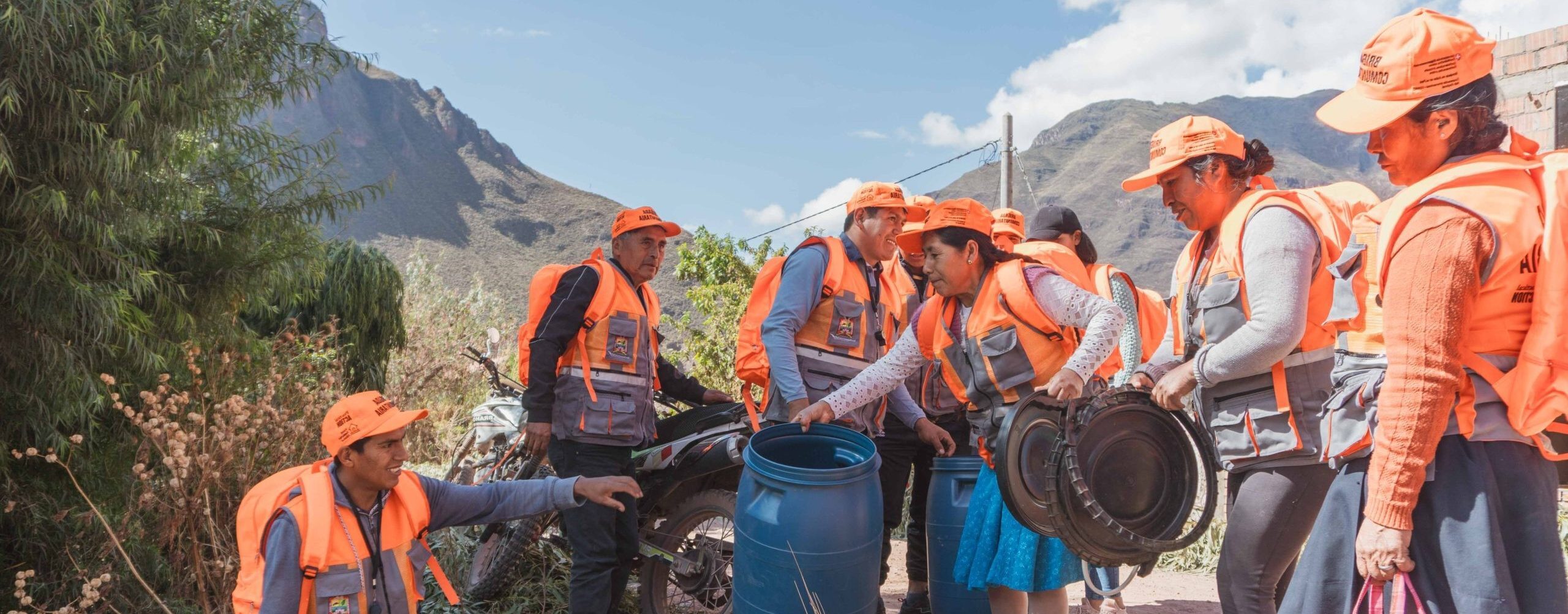 A group of people in orange vests and hats participate in a cleanup activity, collecting trash in large blue barrels outdoors near mountains.