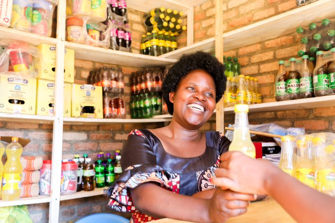 A woman smiles while handing a bottled drink to a customer in a small shop stocked with various beverages.