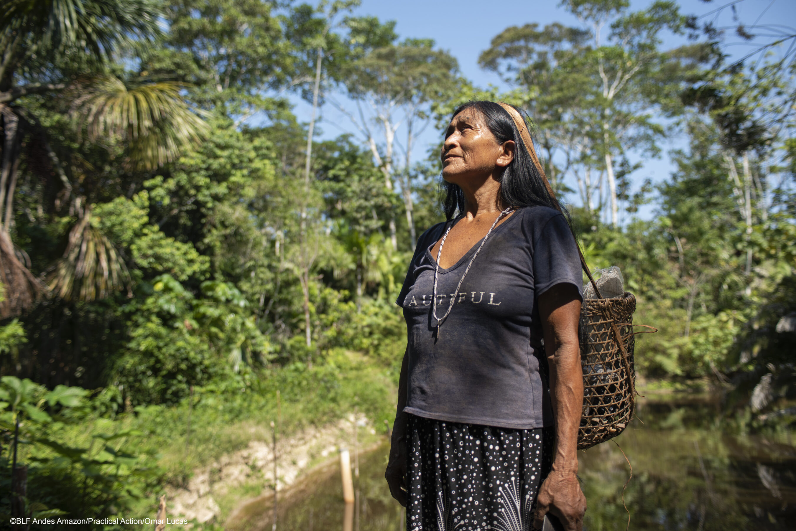 An Indigenous woman stands outdoors in a forested area with lush greenery, wearing a dark shirt and skirt, carrying a woven basket on her back under a sunny sky.