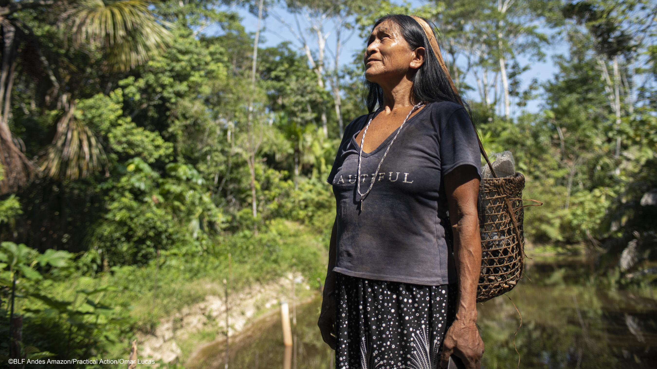 An Indigenous woman stands outdoors in a forested area with lush greenery, wearing a dark shirt and skirt, carrying a woven basket on her back under a sunny sky.