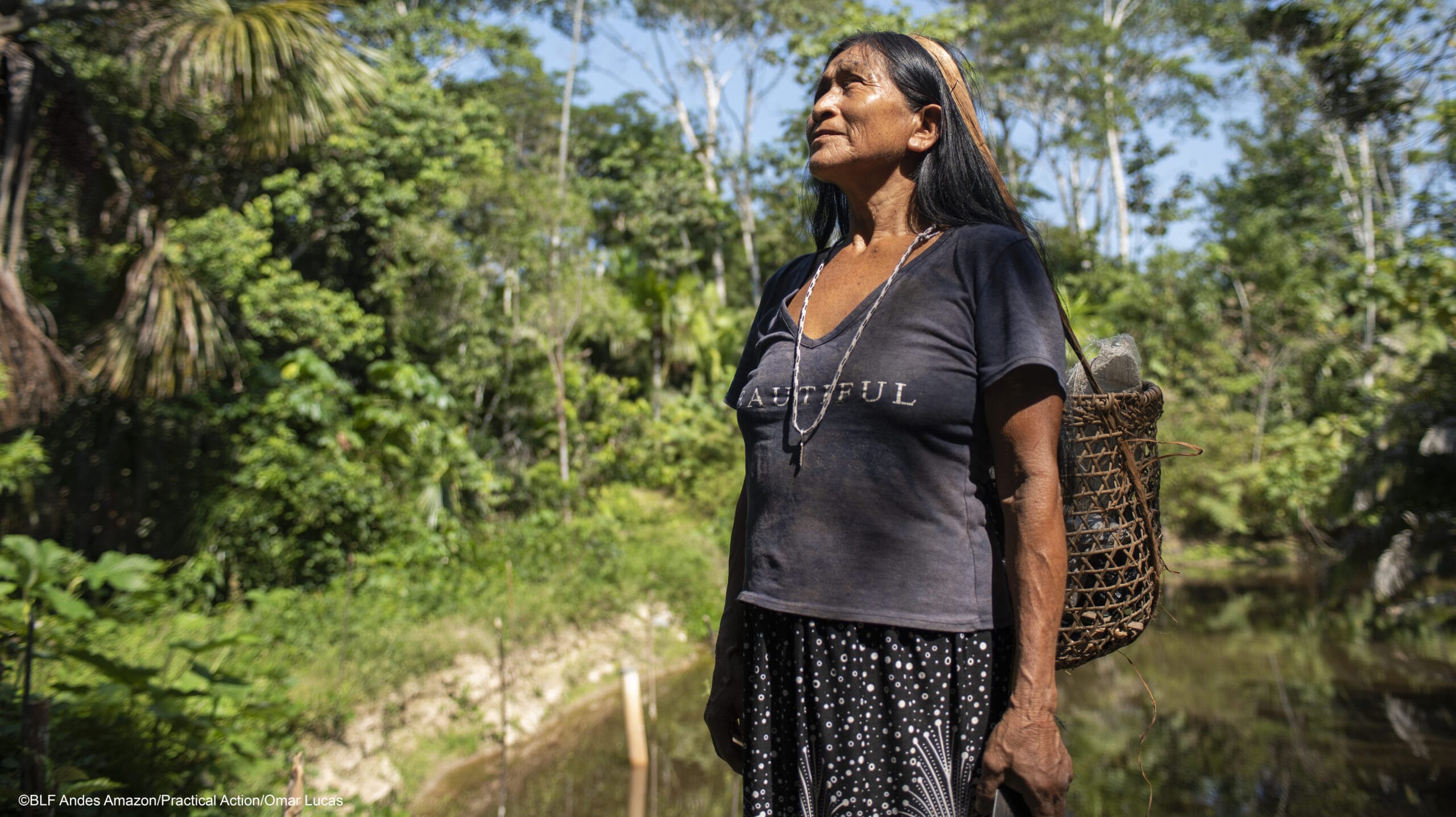 An Indigenous woman stands outdoors in a forested area with lush greenery, wearing a dark shirt and skirt, carrying a woven basket on her back under a sunny sky.