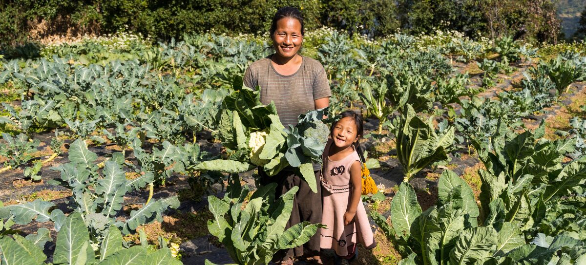 A woman and a young girl stand smiling in a vegetable garden, surrounded by leafy green plants and holding freshly harvested produce.
