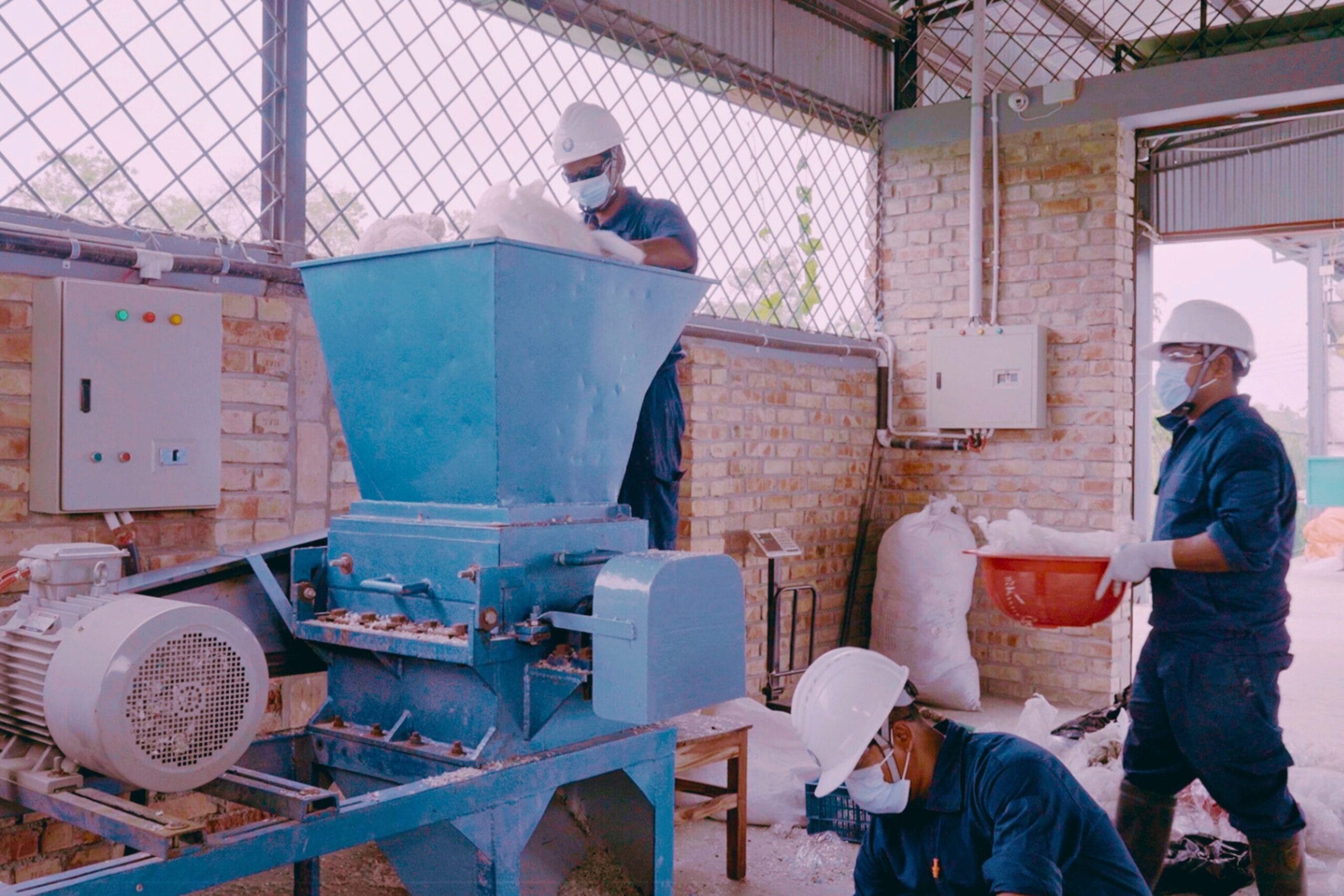 Three workers wearing safety gear operate a blue industrial machine in a factory, handling and processing white material.