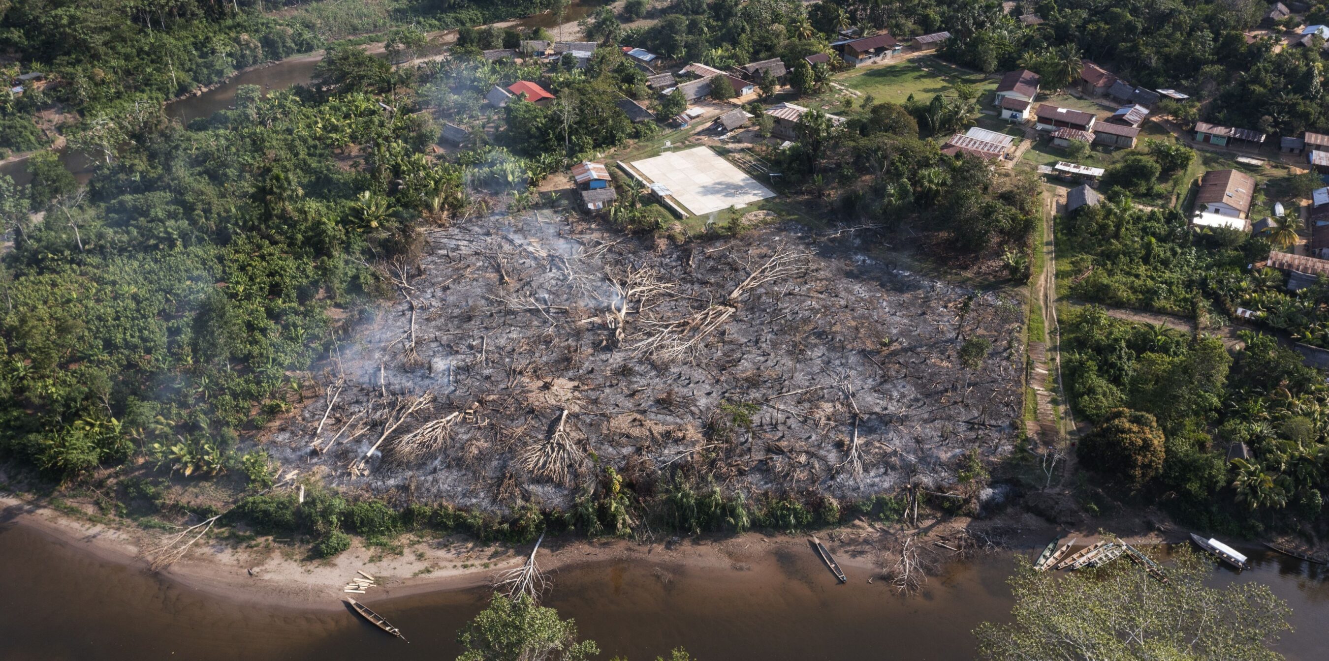 Aerial view of a deforested and burned area near a river, surrounded by dense forest and a small village with several houses and boats.