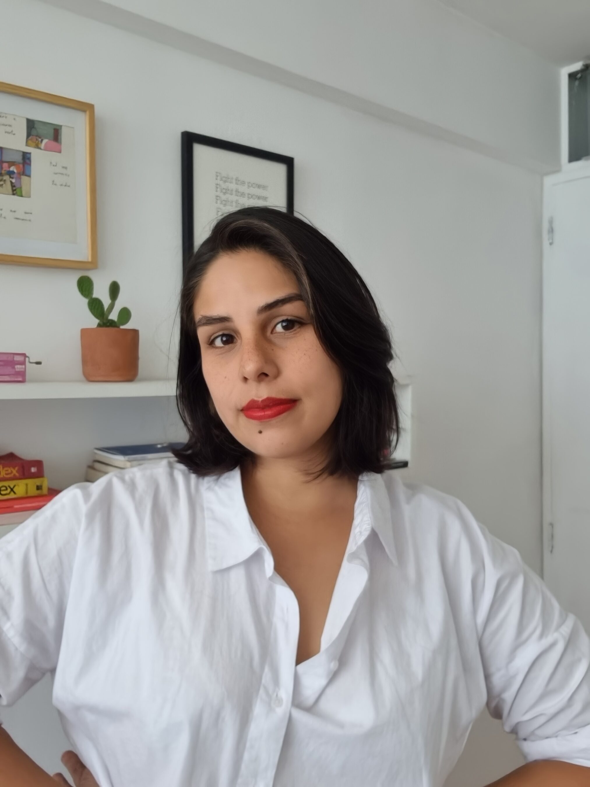 A woman with shoulder-length dark hair and red lipstick stands indoors, wearing a white shirt. Books and a potted cactus are on a shelf in the background.