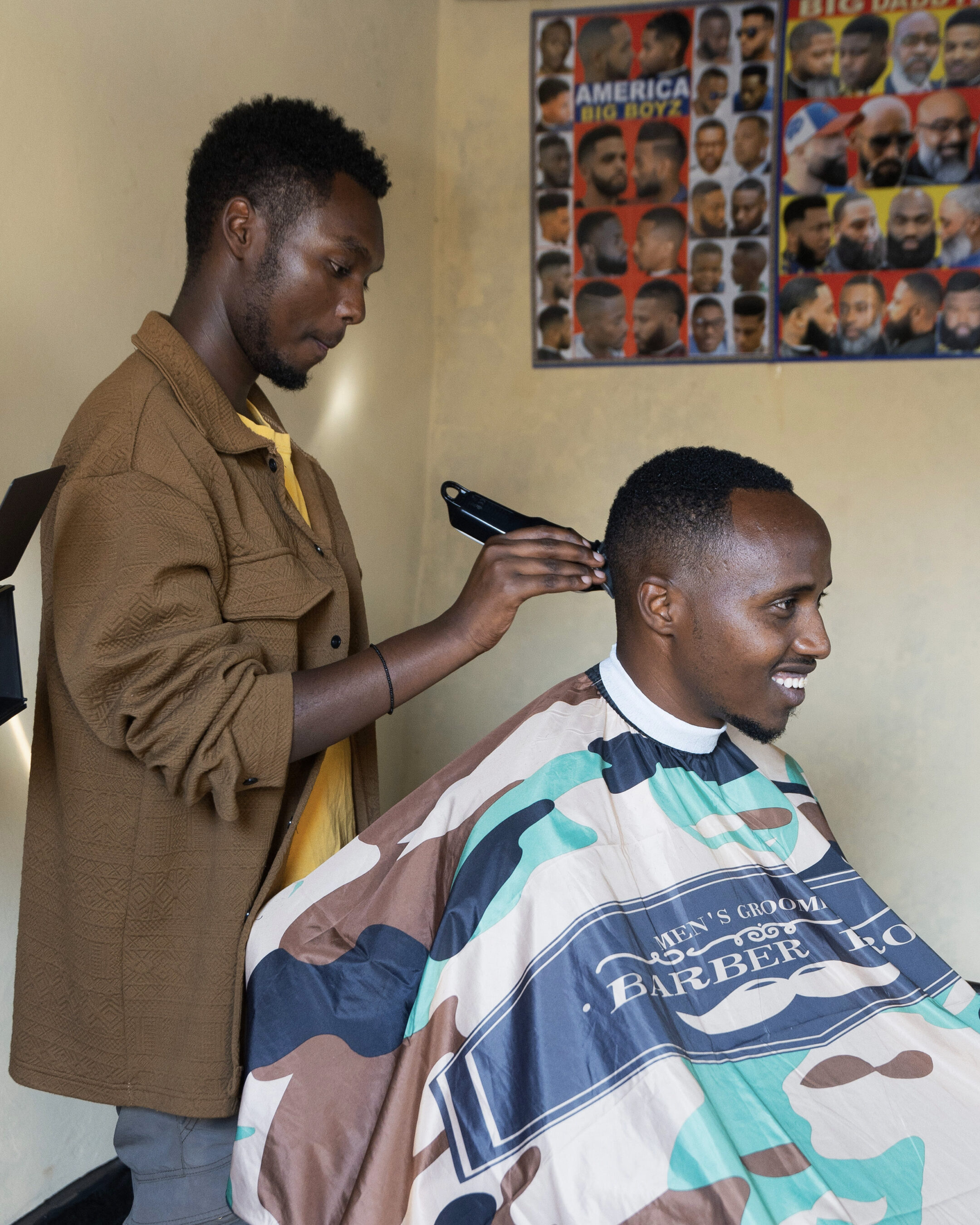 A barber trims a client's hair with clippers in a barbershop. The client wears a camo-patterned cape, and hairstyle posters are visible on the wall.